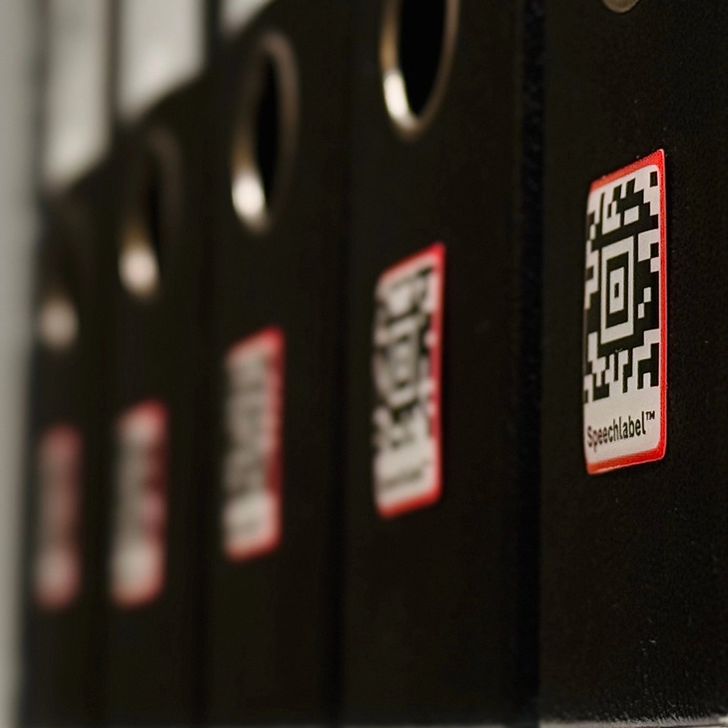 Close-up of a row of black office binders, each marked with a square Speechlabel™ sticker featuring a black-and-white code with a red border. The nearest binder and its Speechlabel sticker are in sharp focus, while the others fade into the background. All labels shown are from Speechlabel, designed for accessible identification with the companion app