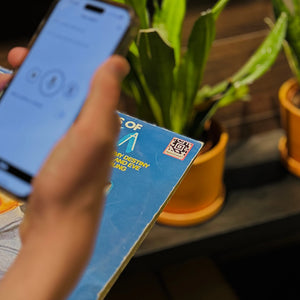 Person holding a phone with a blue screen with the speechlabel app opened. Behind it is an LP with a Speechlabel Epoxy Barcode, plants in the background.