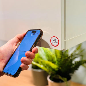 Hand holding a smartphone with a screen displaying a blue interface with the Speechlabel app opened, in front of a NFC sticker from speechlabel product on a white cabinet. The phone is about to tap the label.