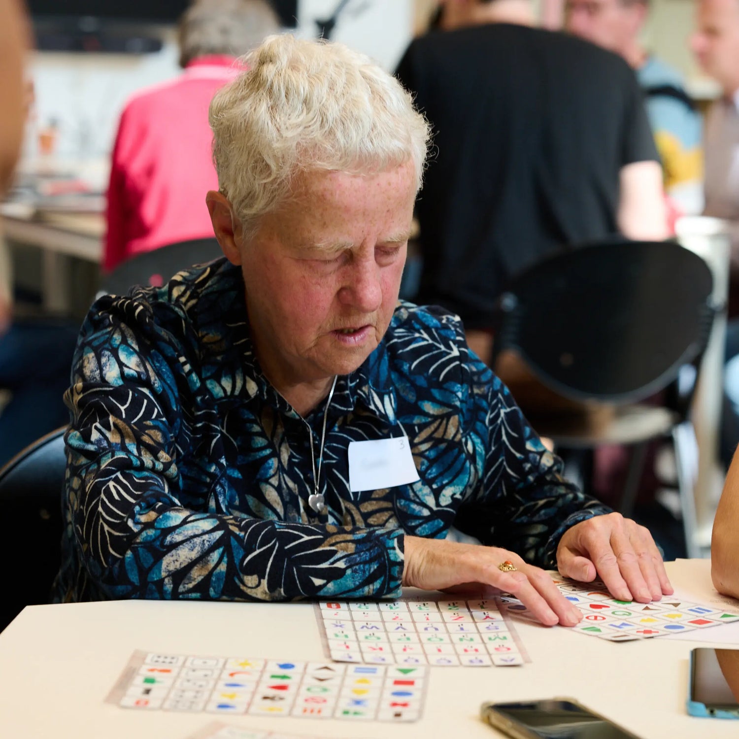 An older woman sits at a table feeling Stactiles tactile sticker sheets with both hands. She appears focused as she explores the raised shapes and symbols on the sheets. Other people are seated and interacting in the background of the room.
