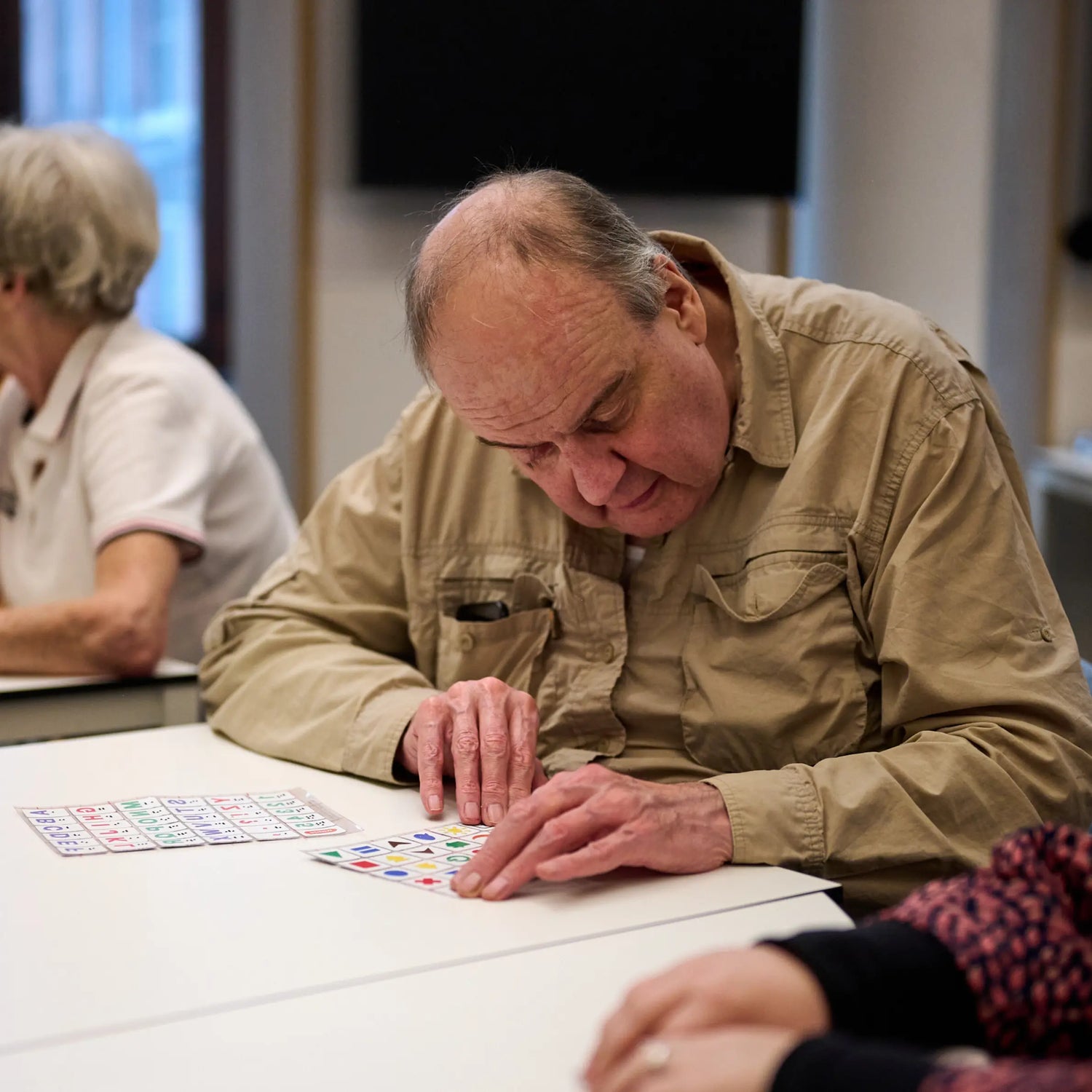 An older man sits at a table exploring Stactiles tactile sticker sheets with his fingertips. He leans in closely as he feels the raised shapes and symbols. Another person is seated beside him in a classroom-like setting.