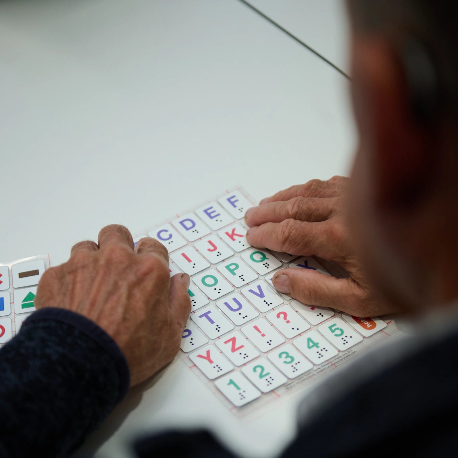 A person uses both hands to feel a Stactiles letters sheet, exploring the raised alphabet stickers arranged in a grid. Another sheet with tactile icons is partially visible beside it on the table.
