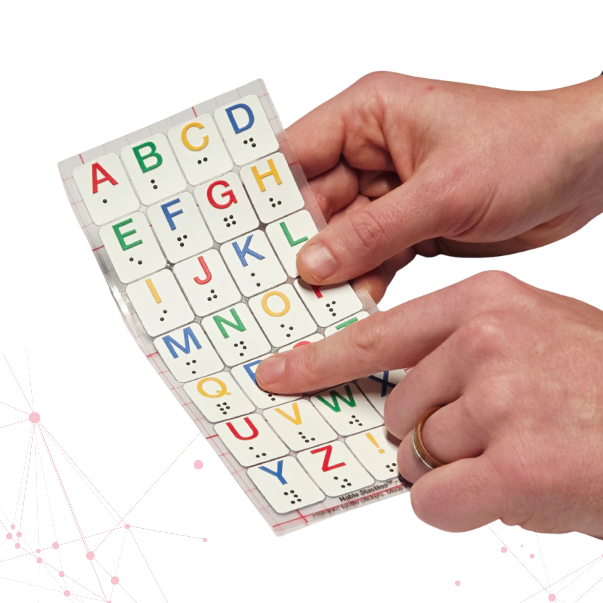 Two hands hold a Stactiles Letters sheet while a finger traces one of the raised, colorful alphabet stickers with braille dots. The tactile letters are arranged in a neat grid on a transparent backing.